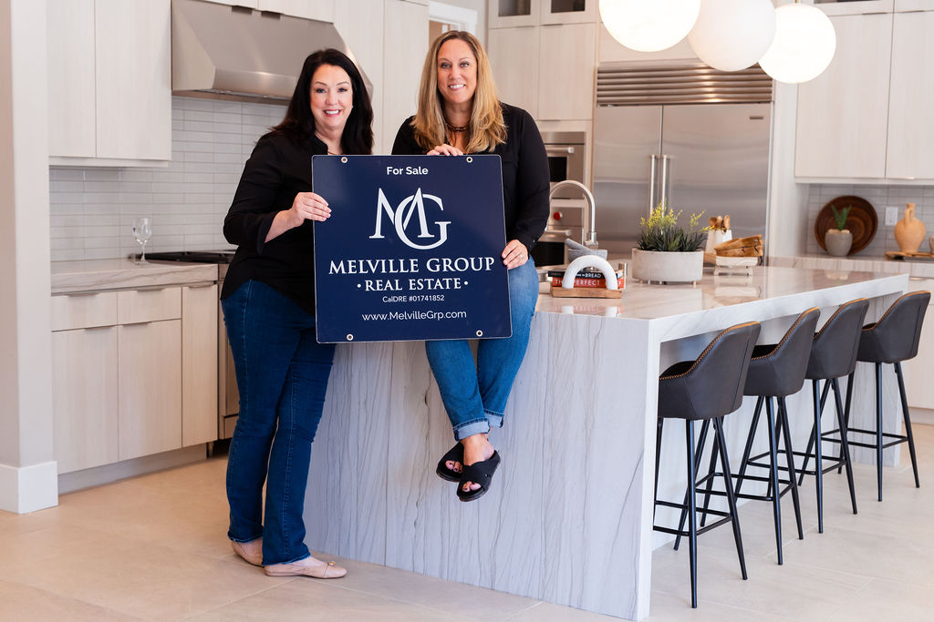 Margaret and Katie, a new partnership,
holding a Melville Group Real estate sign in a beautiful kitchen